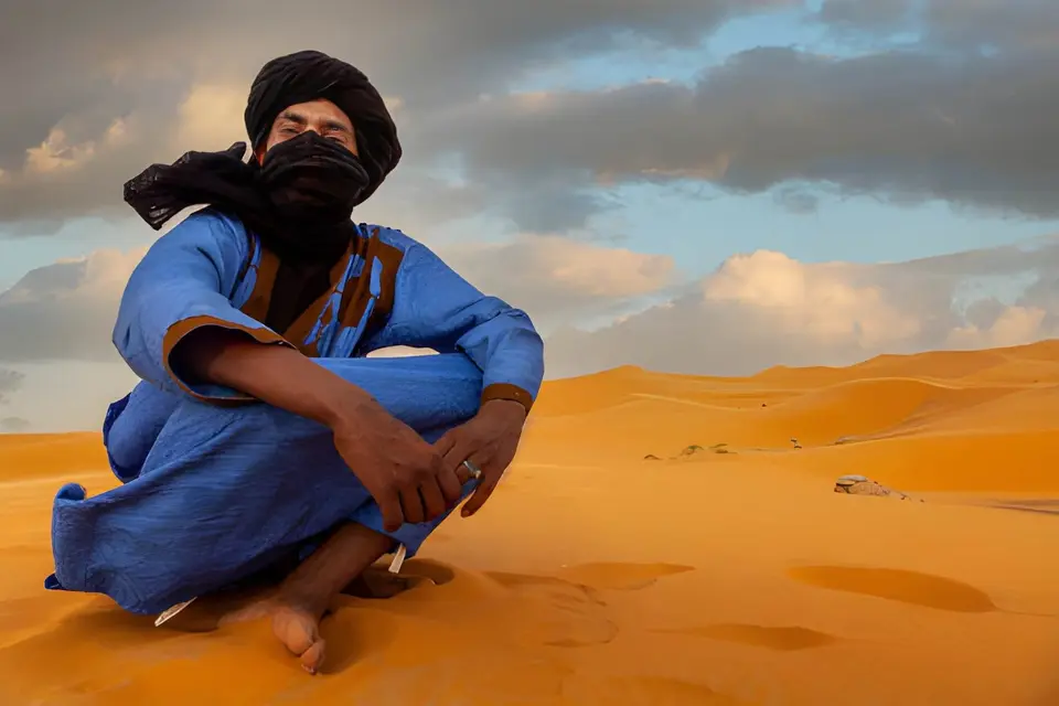 A man in blue robes and a black turban seated on a dune